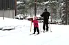 Voksen og barn går på langrenn i snødekt skog ved hus.