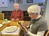 En eldre kvinne ved et bord taster på mobiltelefon. En eldre mann sitter bak henne.