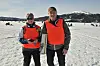 To personer i oransje vester står på islagt vann med folk og fjell i bakgrunnen.