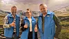 Tre personer i blå vester står foran et fotomotiv av fjell og dal og viser tommel opp.