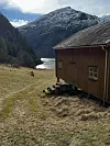 Trehus på gresslette foran fjord og snødekt fjellandskap.