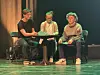Tre unge personer sitter på scenen rundt et lite bord under en teateropptreden.
