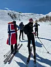 Flere personer går på ski opp en solfylt, snødekt fjellside, én med norsk flagg på ryggen.
