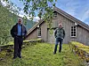 To menn står foran en gammel trebygning med mosegrodd steinmur og grønt gress