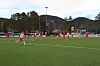 Fotballkamp på kunstgressbane omgitt av trær og åser i bakgrunnen