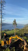 Person styrer en gul minigraver i skogen med fjord og åser i bakgrunnen.