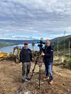 To personer med TV-kamera på stativ ved anleggsområde med fjord og skogkledde åser bak.