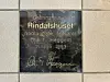 Mørk minneplate i gulv med teksten 'Grunnstein til Rindalshuset nedlagt av ordfører Ola T. Heggem 11. juli 1993' og underskriften til Ola T. Heggem nederst.