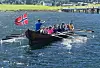 Trebåt full av roere på fjord med norsk flagg og grønt landskap bak