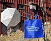 Blått handlenett med tekst foran rød låvevegg og hvit parasoll.