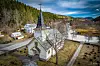Trekirke med høyt tårn ved vei omgitt av gravplass og skogkledde åser.