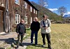 Tre personer foran et gammelt trehus med fjell og blå himmel bak.