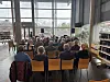 Rundt 30 voksne mennesker sitter på stoler vendt mot en dame i et lyst bibliotek.