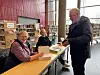 Forfatter signerer bok for en besøkende ved et bord inne på et bibliotek.