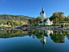 Hvit kirke ved fjordkanten speiler seg i stille vann omgitt av åser og trær
