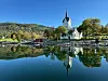 Hvit kirke ved fjordkanten speiler seg i stille vann omgitt av åser og trær