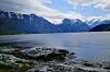 Steinete strand ved stille fjord omgitt av snødekte fjell under skyet himmel