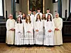 Konfirmanter i hvite kapper poserer med to prester foran alteret i en kirke.
