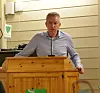 Mann står bak trett talerstol og snakker i mikrofon innendørs.