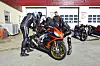 Motorsyklister i skinndresser står rundt en oransje sportssykkel foran et verksted.
