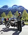 Motorsyklist står ved motorsykkel foran snødekt fjell og grønne trær