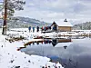 Snødekt landskap med en liten bygning ved vannet og mennesker som går langs bredden.
