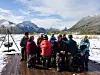 Gruppe turister sitter på benk ved en snødekt innsjø med fjell i bakgrunnen.
