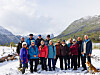 En gruppe turkledde folk og en hund står sammen i snøen foran fjell og vann under en lys himmel.