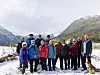 En gruppe turkledde folk og en hund står sammen i snøen foran fjell og vann under en lys himmel.