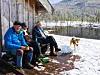 To personer sitter ved en trekledd hytte, med en hund stående i snøen foran dem.