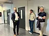 Tre personer står i en korridor og snakker, med dører og plakater i bakgrunnen.