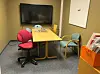 Lite møterom med bord, flere stoler, veggmontert skjerm og en orange dokumenthylle.