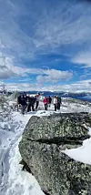 Turgåere går på en snødekt fjellsti med stor stein i forgrunnen og vid utsikt mot fjell.