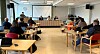 Møterom med publikum ved bord og panel ved langpult foran prosjektorlerret.