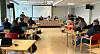Møterom med publikum ved bord og panel ved langpult foran prosjektorlerret.