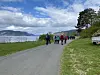 Turgåere på en vei ved vannet med fjell, trær og skyet himmel.