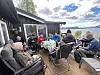 Flere personer sitter rundt et bord på en terrasse med utsikt over et vann og fjell i bakgrunnen.