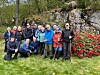 En gruppe turkledde mennesker står og sitter samlet på gresset ved en steinbakke med røde blomster.