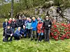 En gruppe turkledde mennesker står og sitter samlet på gresset ved en steinbakke med røde blomster.