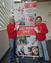 To personer i røde gensere står ved et kampanjebanner og holder et skilt med teksten «Bli med oss!».