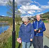 To personer i blå jakker står ved en sti nær vannet med landskap og hus i bakgrunnen.