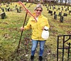 Person i gul jakke holder en rive og en sprayflaske på en kirkegård med mange gravsteiner.