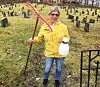 Person i gul jakke holder en rive og en sprayflaske på en kirkegård med mange gravsteiner.