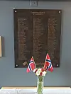 Minneplate på veggen med to norske flagg og en blomsterbukett i en vase på et bord.