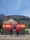Tre personer står på et podium med to spanske flagg og et norsk flagg foran et fjell og en kirketårnspire.