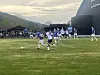Spillere i blå og hvite drakter kjemper om ballen på en fotballbane med tribune og fjell bak.