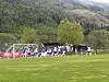 Fotballspillere samles foran mål på en gressbane med skogkledde fjell i bakgrunnen.