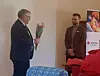 To menn står i et lyst rom med blomster, sofa og Røde Kors-plakat i bakgrunnen.