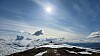 Solen lyser over en snødekt fjellrygg med skyer og fjell i horisonten.