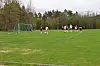 Flere fotballspillere på en grønn bane med mål og skog i bakgrunnen.
